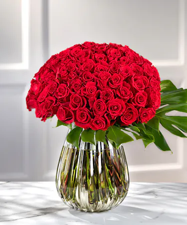 100 Luxury Red Roses artfully designed in a romantic, bloom-on-bloom presentation with exotic tropical foliage in our teardrop decor vase.